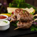 Grilled Lamb Kofta kebabs on wooden skewers served with creamy mint yogurt sauce and fresh parsley on a platter.
