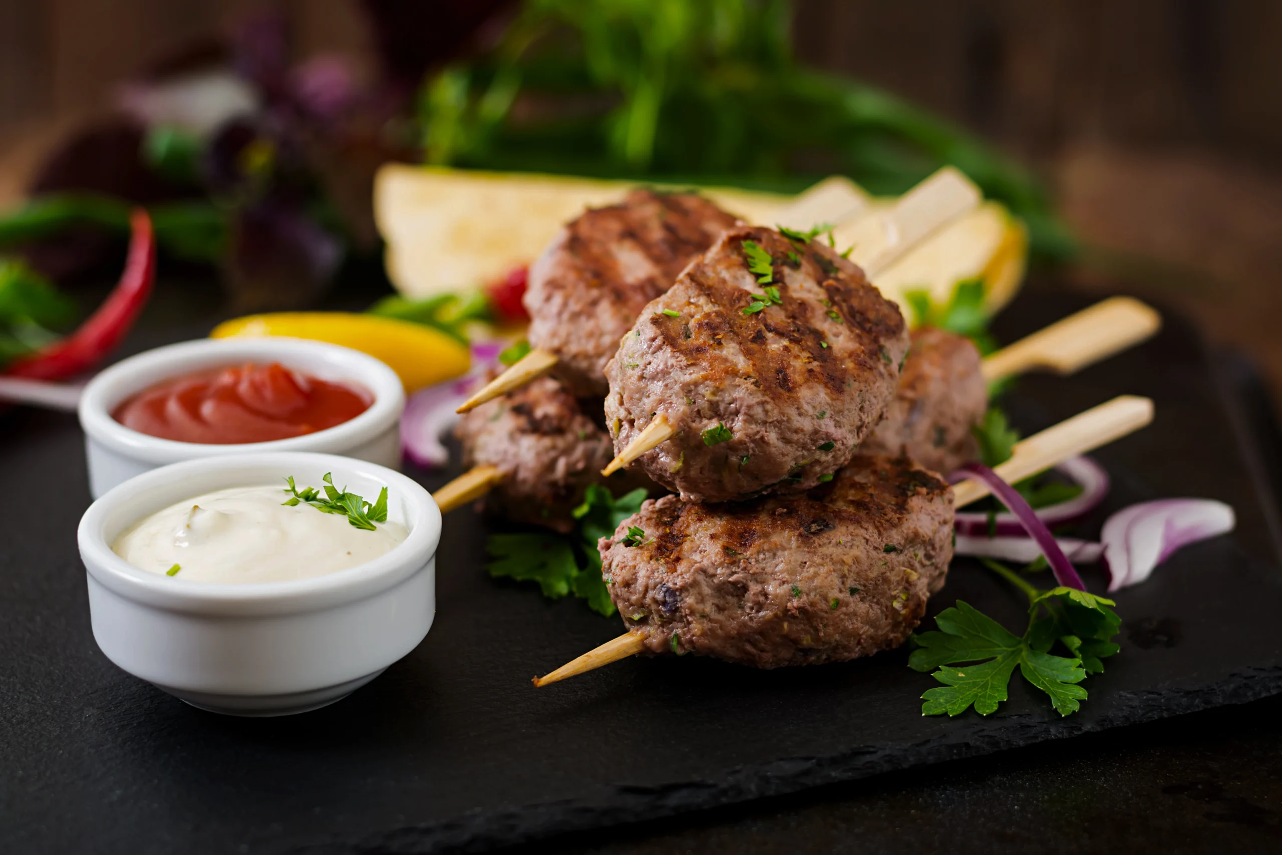 Grilled Lamb Kofta kebabs on wooden skewers served with creamy mint yogurt sauce and fresh parsley on a platter.