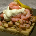 A gourmet prawn mayonnaise sandwich on thick-cut malted bloomer bread with fresh baby gem lettuce and a light dusting of smoked paprika on a rustic plate.