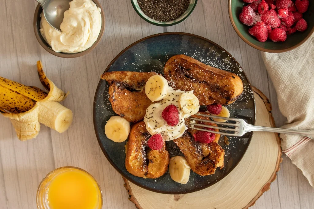 Classic golden French toast served on a white plate with crispy bacon strips and a side of fresh seasonal fruit