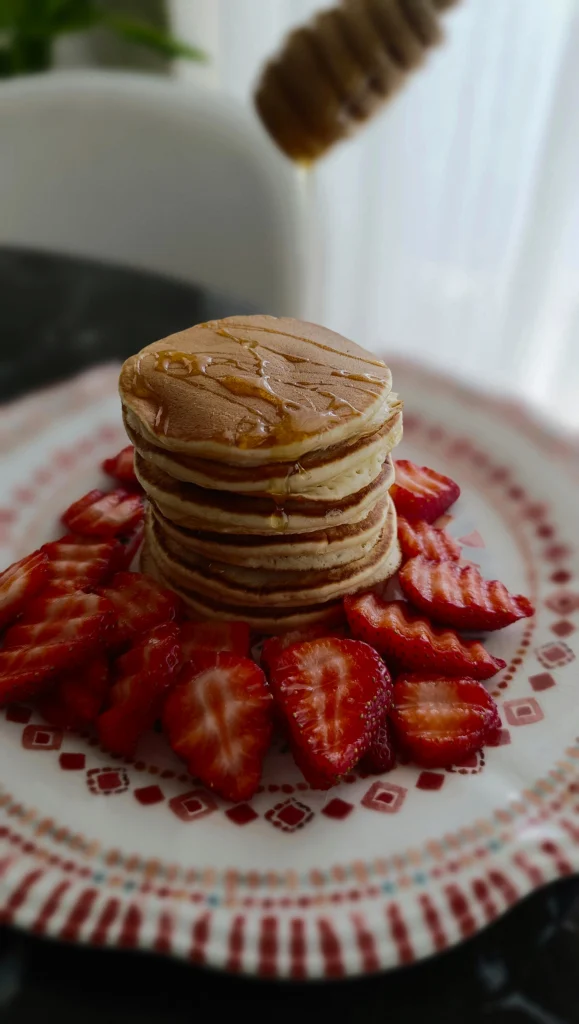 A tall stack of fluffy Scotch pancakes served on a white plate, topped with melting butter and a drizzle of syrup, showcasing a golden-brown texture.