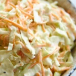 A shallow bowl filled with classic coleslaw featuring shredded green cabbage and carrots tossed in a creamy dressing, sprinkled with black pepper.